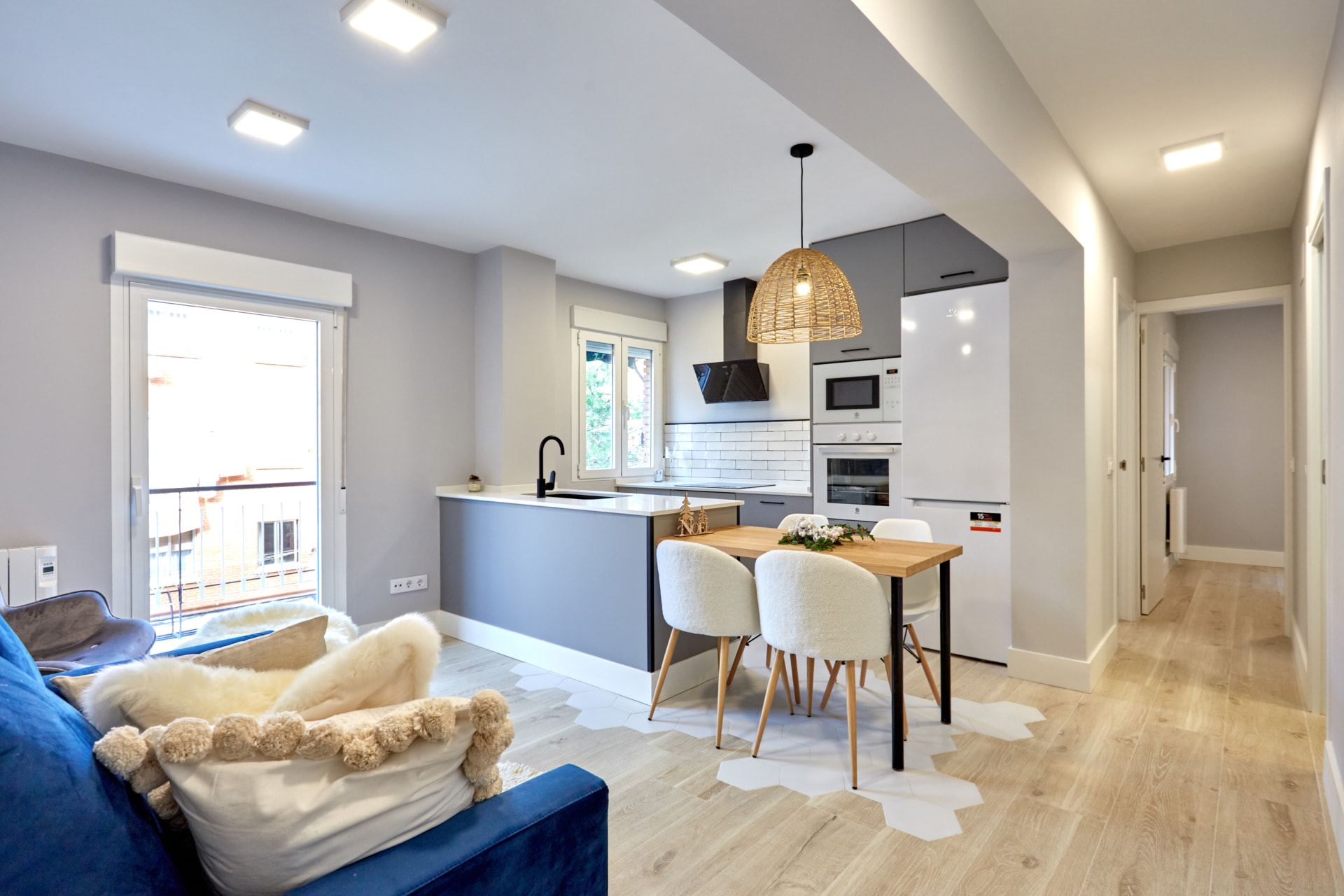 Salón-cocina diáfano con sofá azul, mesa de madera, sillas blancas y lámpara de ratán