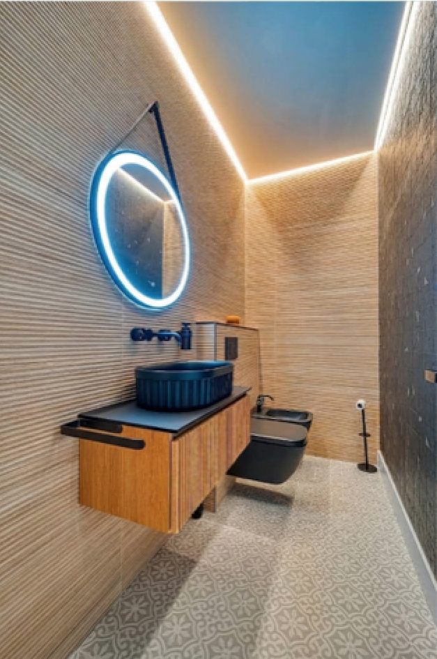 Aseo con azulejos beige texturizados, lavabo azul oscuro, mueble de madera y espejo circular iluminado